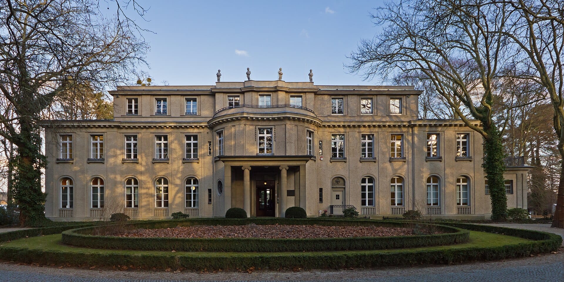 Vista frontale della Casa della Conferenza di Wannsee, una storica villa color beige con finestre simmetriche e giardini curati in una giornata serena.
