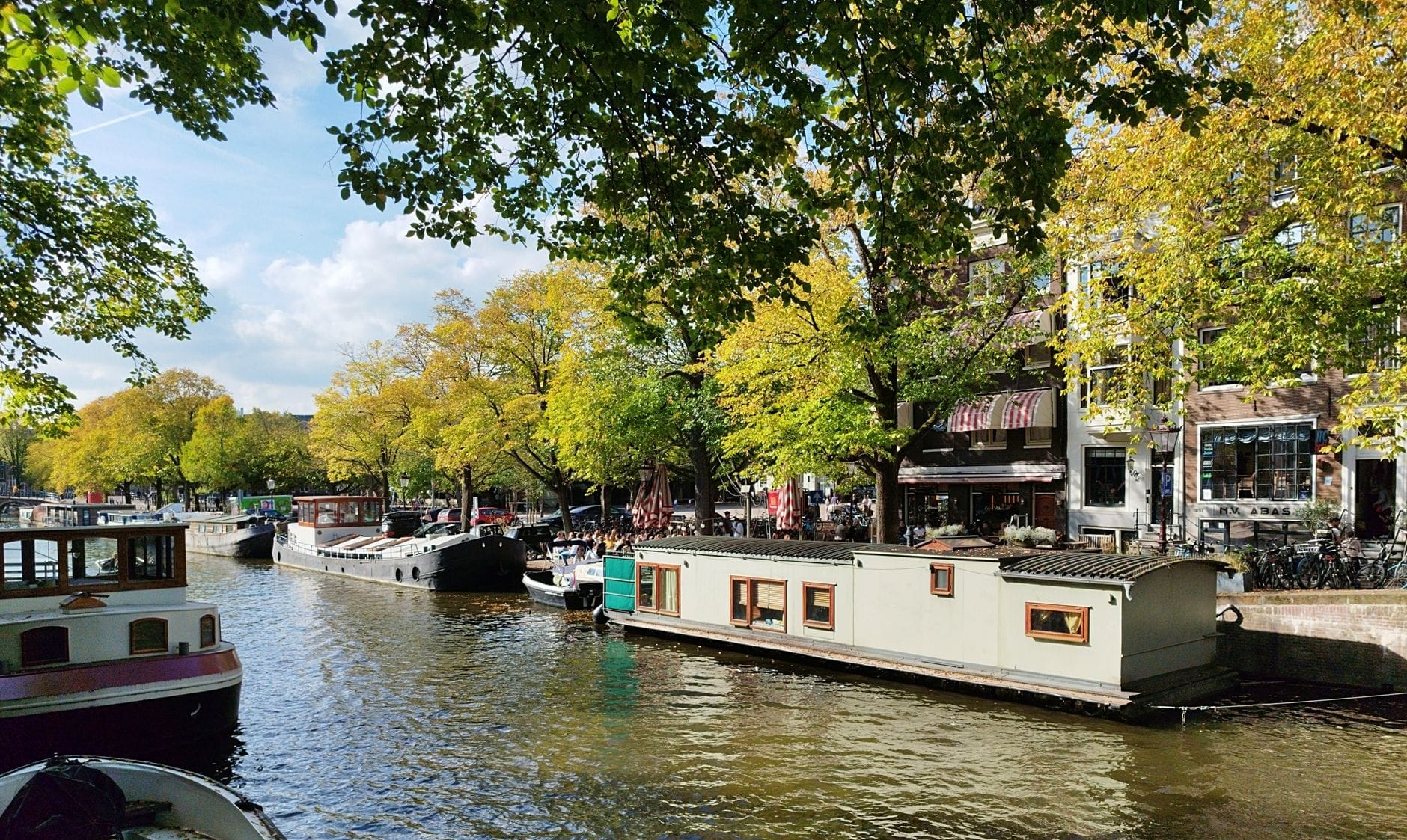 Una hilera de coloridas casas flotantes amarradas a lo largo de un canal de Ámsterdam bordeado de frondosos árboles y edificios históricos de ladrillo en un día soleado.