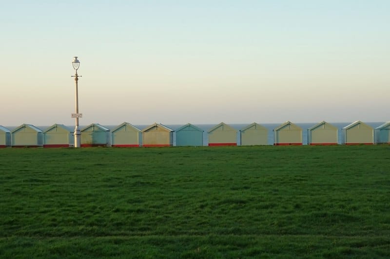 Row of pastel beach huts and a lamp post along the green lawns at Hove Lawns, with the sea visible under a clear sky.