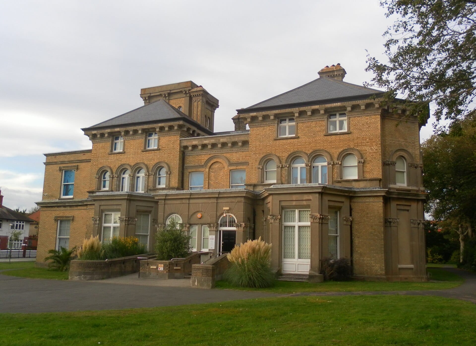 The exterior of Hove Museum of Creativity, a grand Victorian villa with ornate brickwork, surrounded by lawns and greenery under an overcast sky.