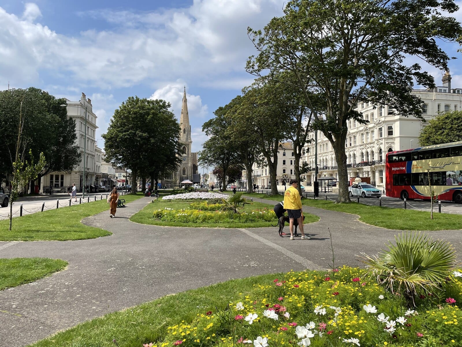Wide boulevard in Hove with flower beds, mature trees, historic white buildings, a church steeple, and a red Brighton & Hove bus under blue sky.