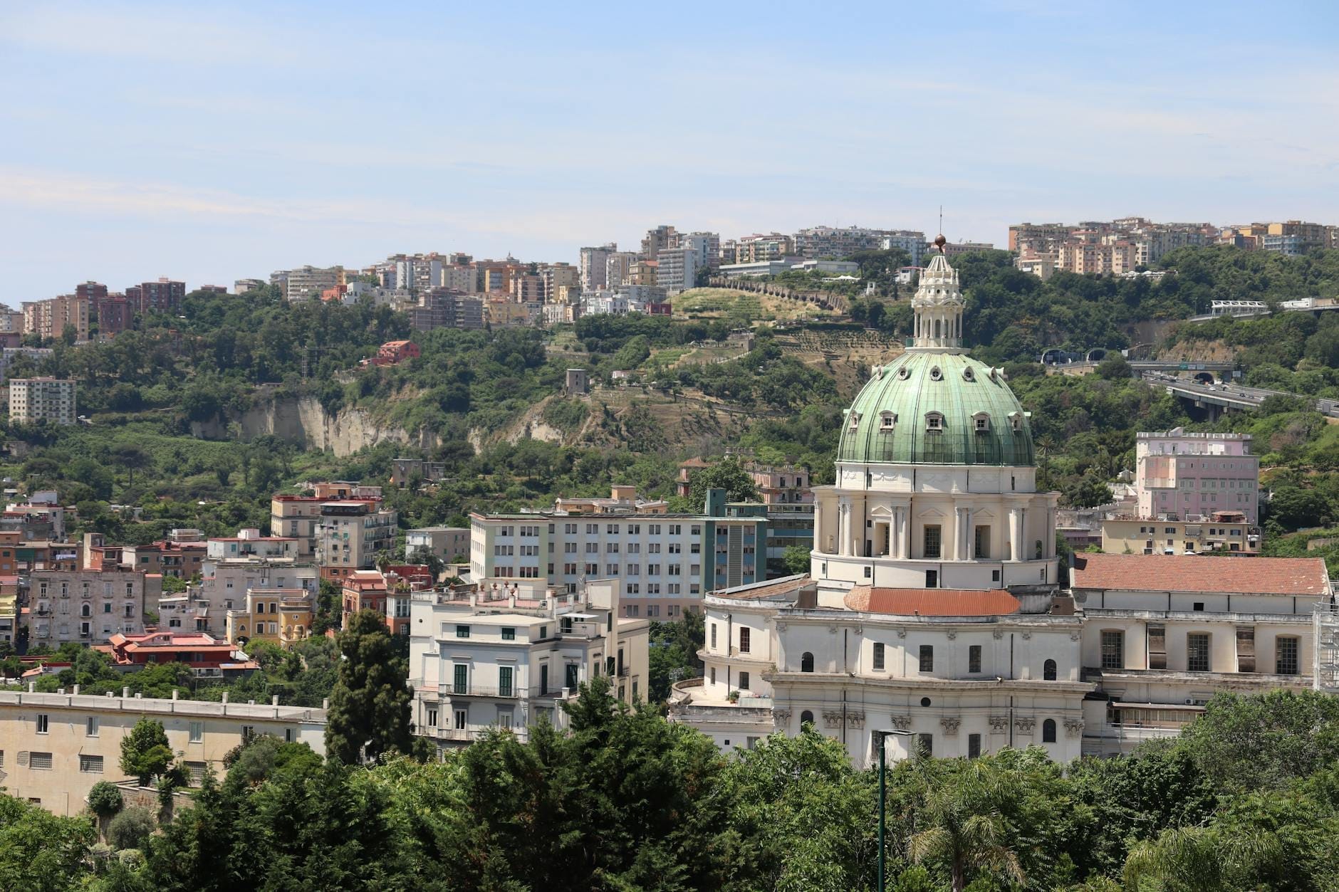 Bâtiment à coupole entouré de verdure en périphérie de Naples avec des collines et un paysage urbain en arrière-plan sous un ciel lumineux.