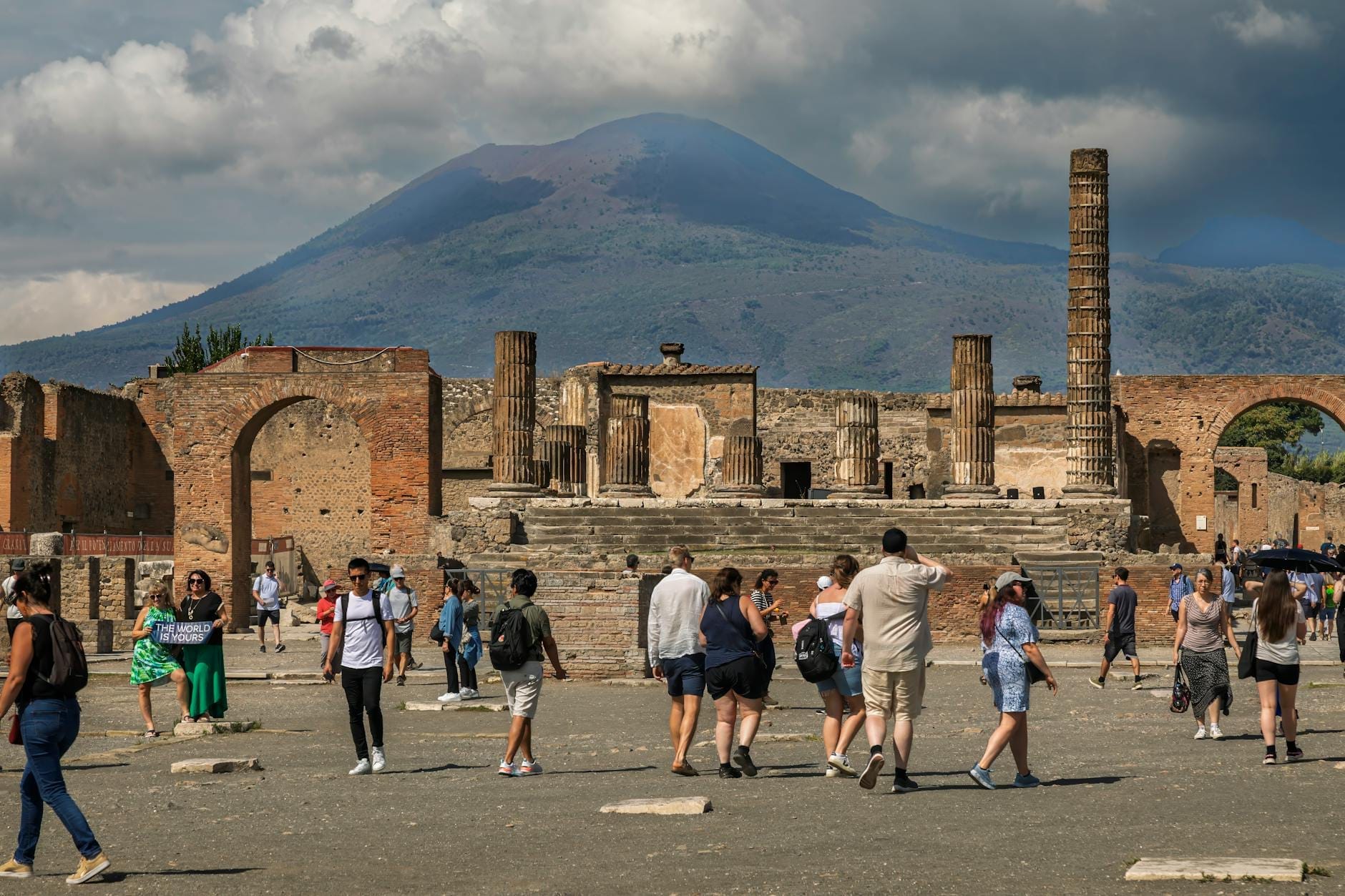 Des touristes se promènent dans les ruines antiques de Pompeii avec le Mont Vésuve en arrière-plan sous un ciel partiellement nuageux.