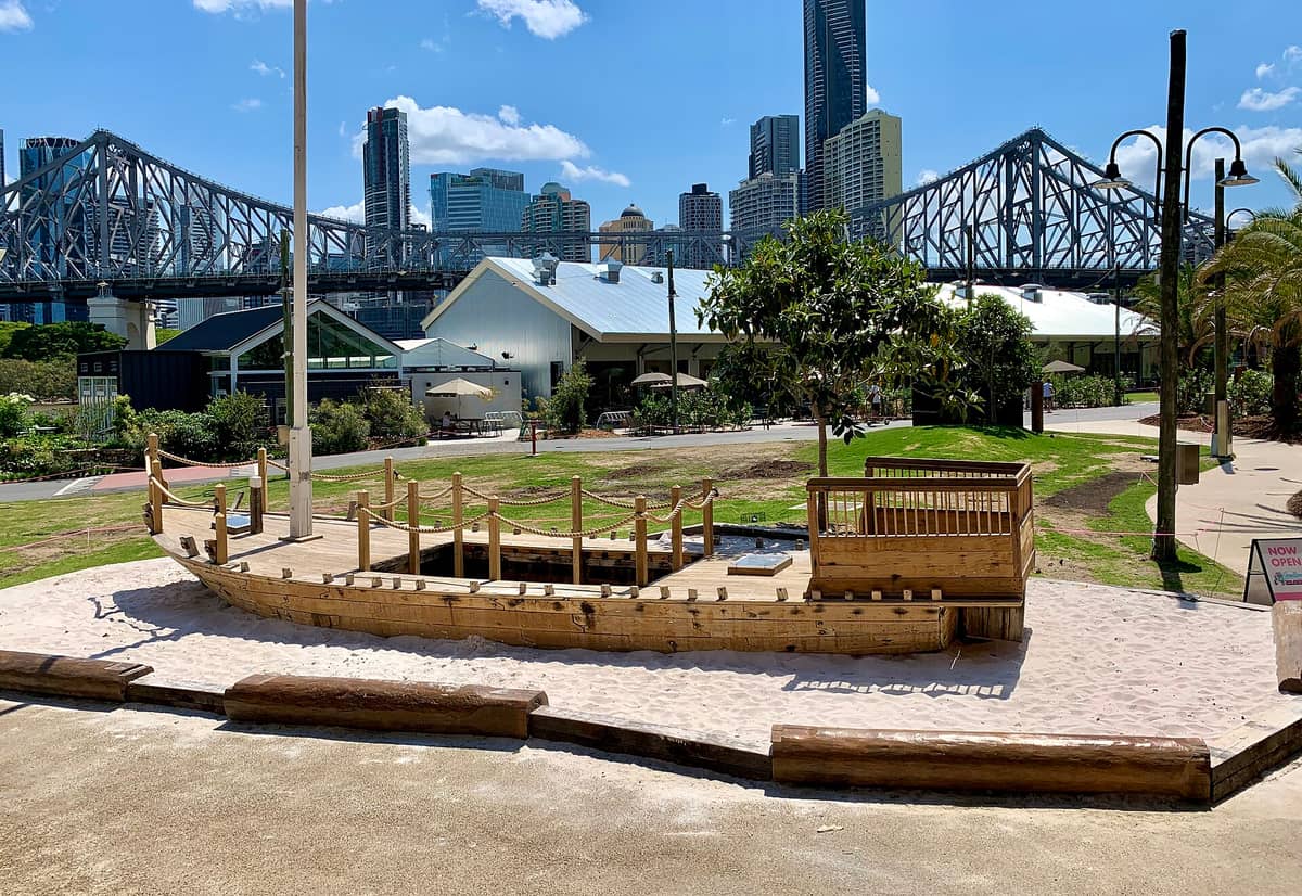 Howard Smith Wharves precinct showing heritage warehouses, Story Bridge, Brisbane city skyline, and playground area under a sunny blue sky.
