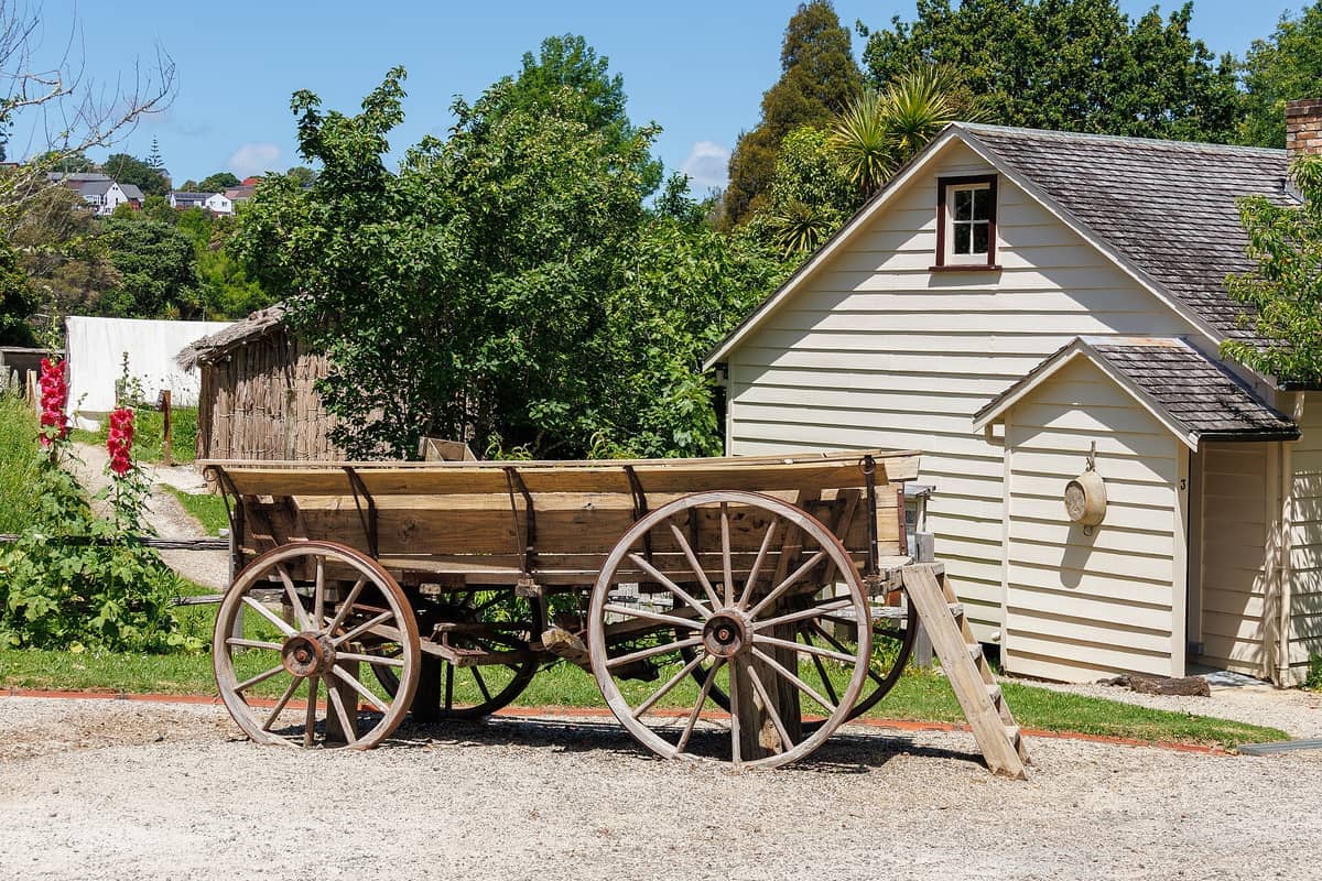 Ein rustikaler Holzwagen steht neben einem cremefarbenen Kolonialhaus im Howick Historical Village an einem sonnigen Tag in Auckland.