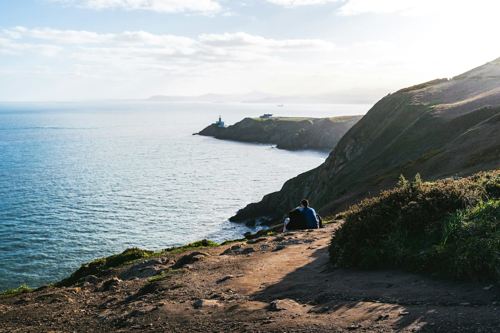 Due persone sedute lungo il sentiero del Howth Cliff Walk, con vista sulle scogliere, il Mare d'Irlanda e un faro lontano sotto un cielo sereno.