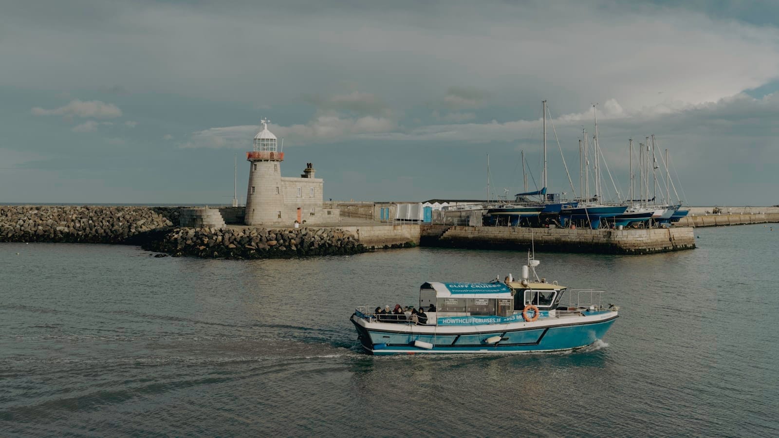 Un peschereccio passa davanti all'iconico faro del porto di Howth, con yacht e il frangiflutti sullo sfondo sotto un cielo luminoso e nuvoloso.