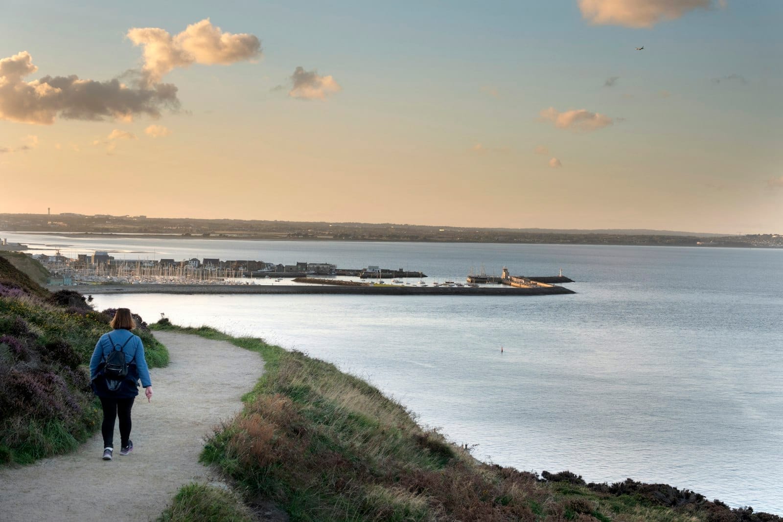 คนหนึ่งเดินตามทางขอบหน้าผา มองเห็นท่าเรือ Howth, มาริน่า, และประภาคารท่ามกลางพระอาทิตย์ตก ท้องฟ้าสีจัดจ้านและวิวริมทะเล