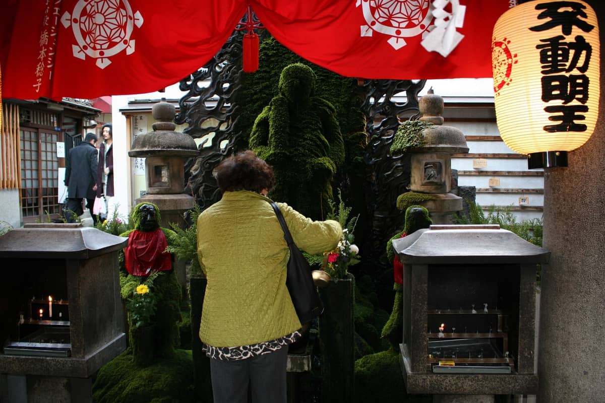 A visitor in a green jacket offers prayers before the moss-covered Fudō Myōō statue at Hozenji Temple, flanked by traditional lanterns and shrines.