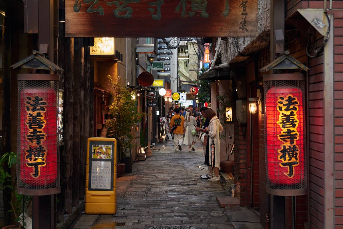 Entrance to Hozenji Yokocho in Osaka with stone-paved lane, traditional lanterns, and people walking between cozy restaurants and bars at dusk.