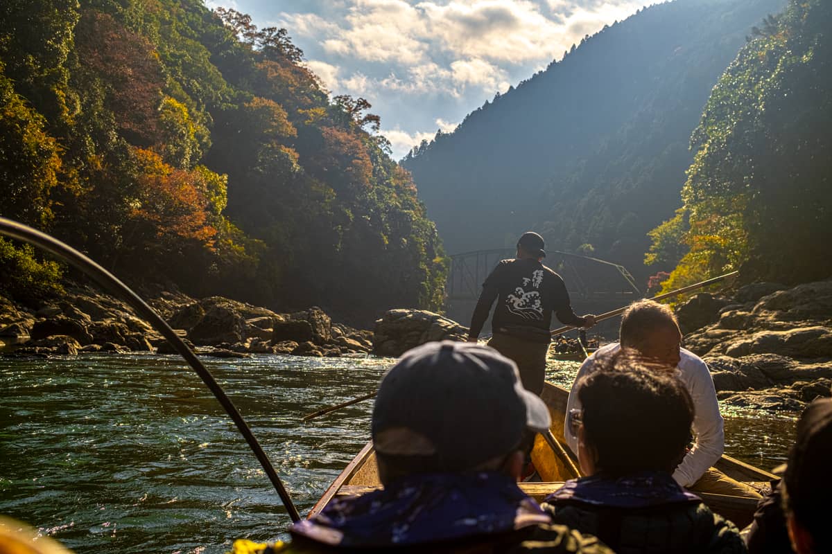 View from inside a traditional wooden boat as it travels down the Hozu River gorge in Kyoto, with passengers, boatmen, and forested mountains on both sides.