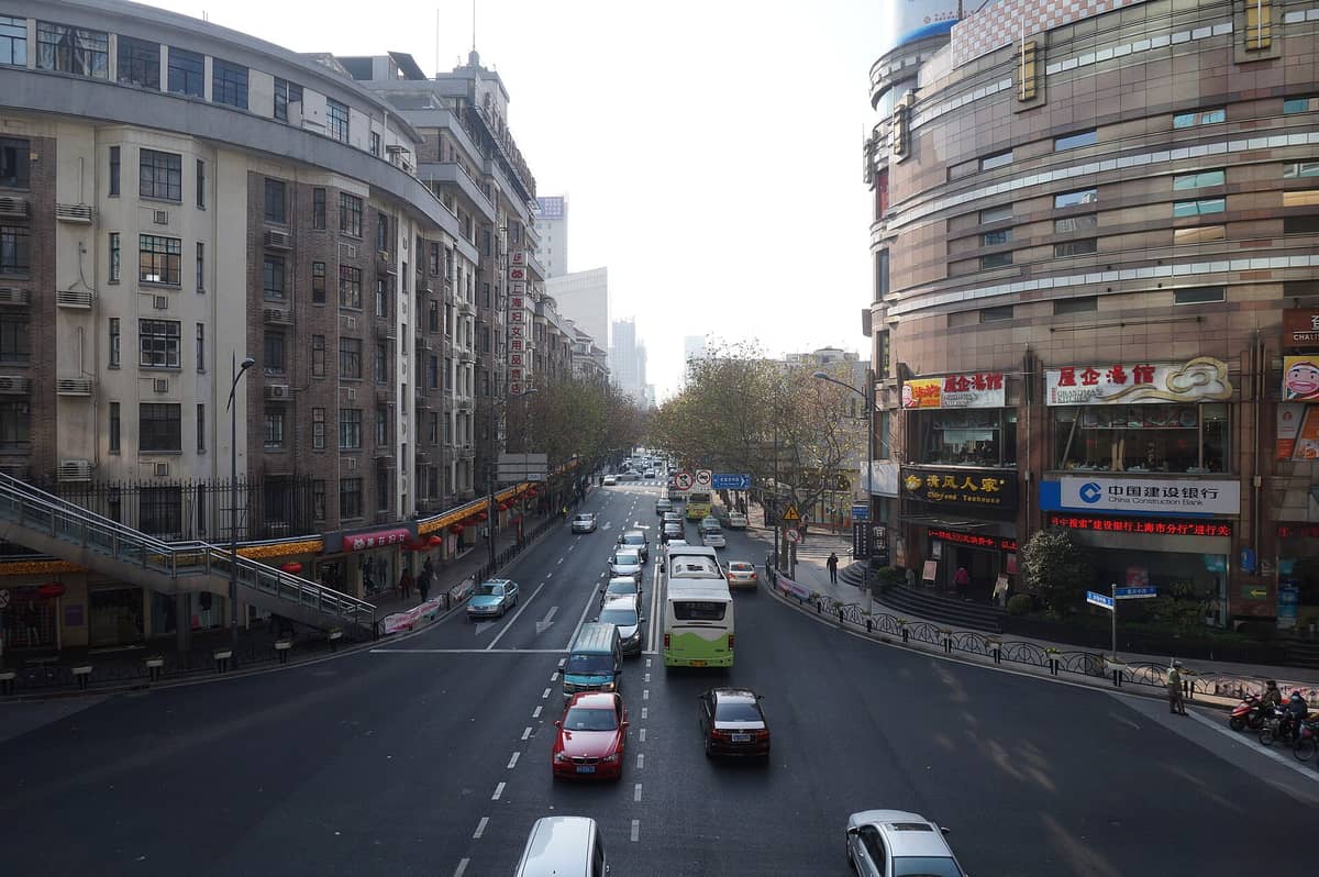 Tageslichtaufnahme der Huaihai Middle Road in Shanghai aus erhöhter Perspektive: baumgesäumte Straßen, historische Gebäude mittlerer Höhe, Ladengeschäfte und lebhafter Straßenverkehr.