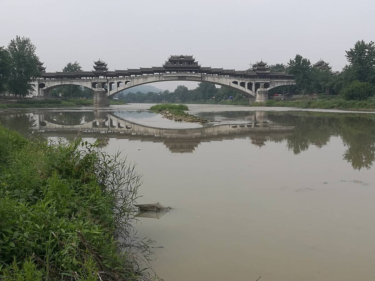 ภาพสะพานไม้โบราณสไตล์จีนข้ามแม่น้ำในเมืองโบราณหวงหลงซี รายล้อมด้วยต้นไม้และเงาสะท้อนในน้ำ