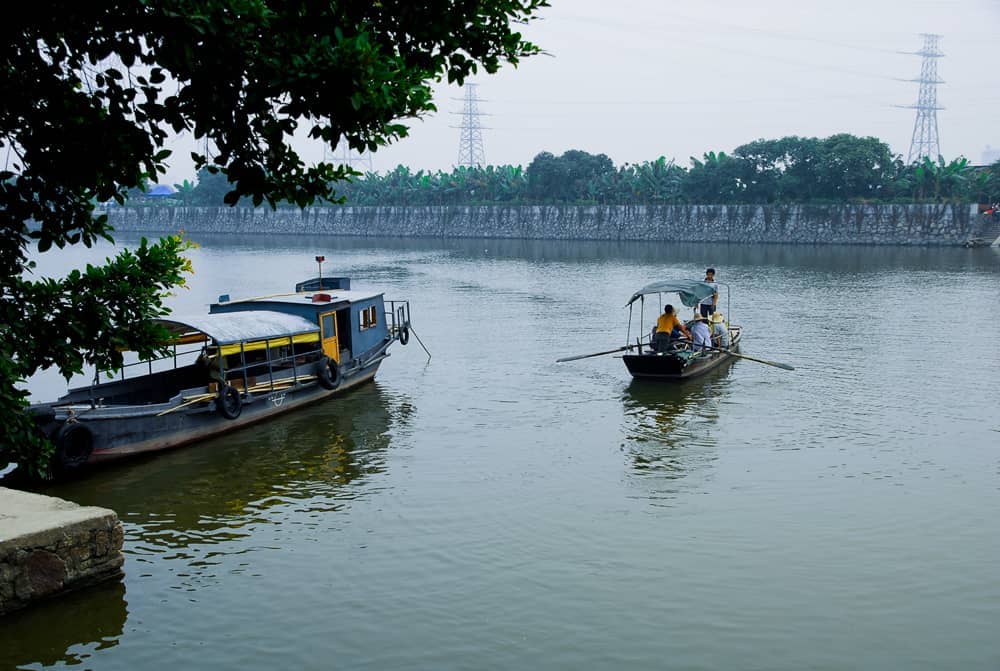 Traditional boats float on the calm river at Huangpu Ancient Port, with leafy trees framing the historic waterfront and stone quay.
