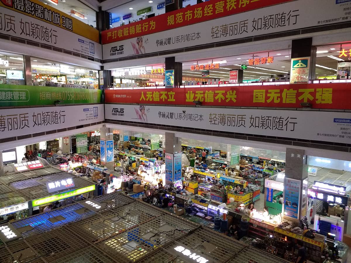 Vista interior do Mercado de Eletrônicos Huaqiangbei em Shenzhen, mostrando vários andares repletos de bancas de componentes eletrônicos e letreiros coloridos.