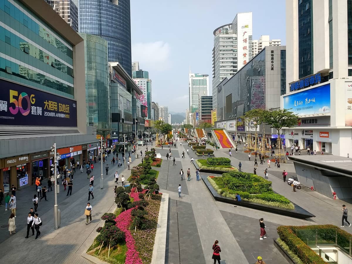Ampla rua pedestre de Huaqiangbei ladeada por shoppings de eletrônicos, lojas de varejo e multidões de pessoas, com arranha-céus ao fundo.