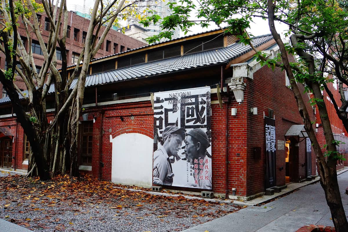 Außenansicht eines roten Backsteingebäudes mit traditionellem Ziegeldach im Huashan 1914 Kreativpark, geschmückt mit großen Postern und umgeben von Bäumen mit Herbstlaub.