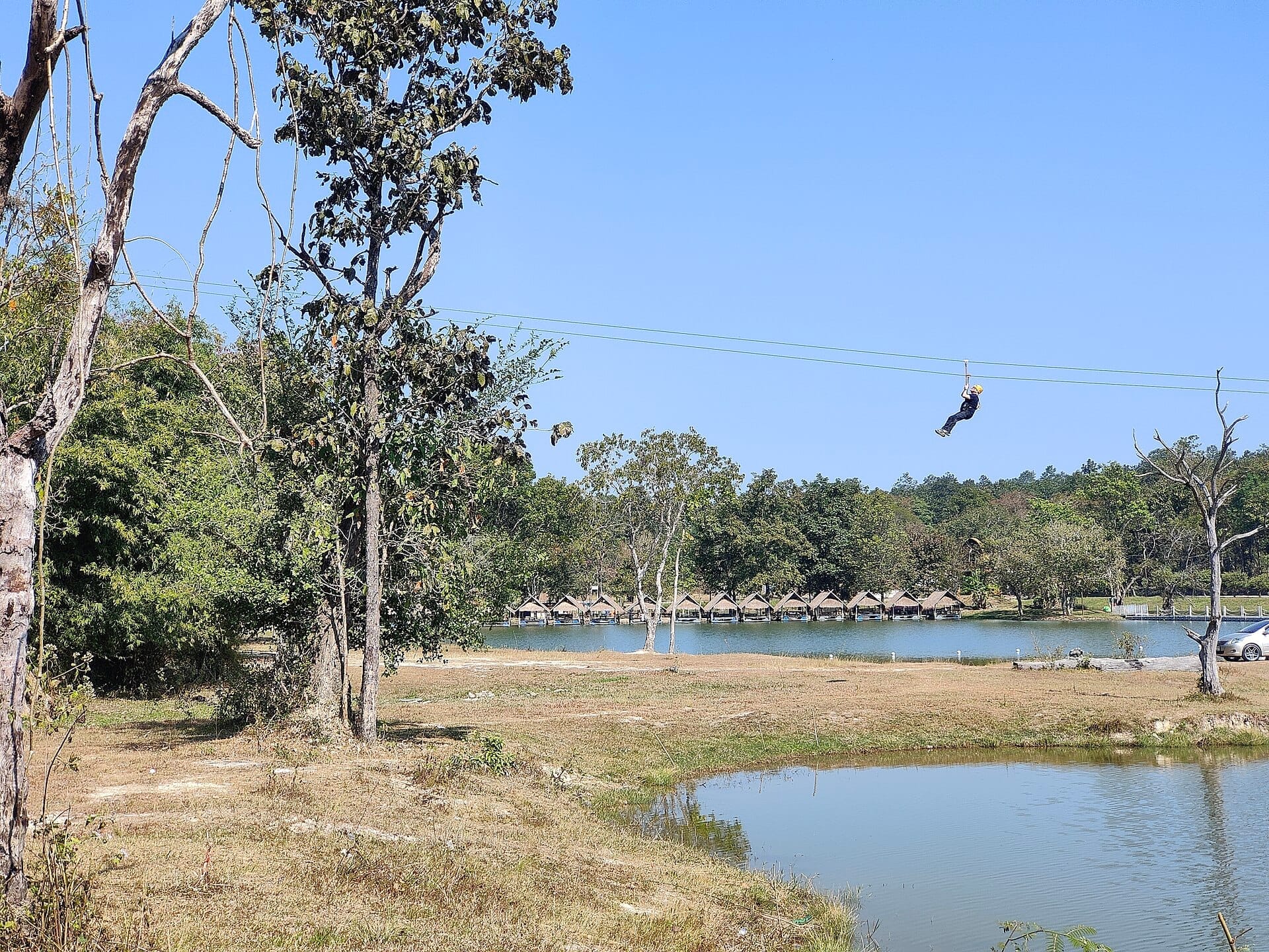 Một người đang chơi zipline trên hồ Huay Tung Tao, với những căn lều tre ven hồ, cây cối và bầu trời xanh dưới ánh nắng buổi trưa.
