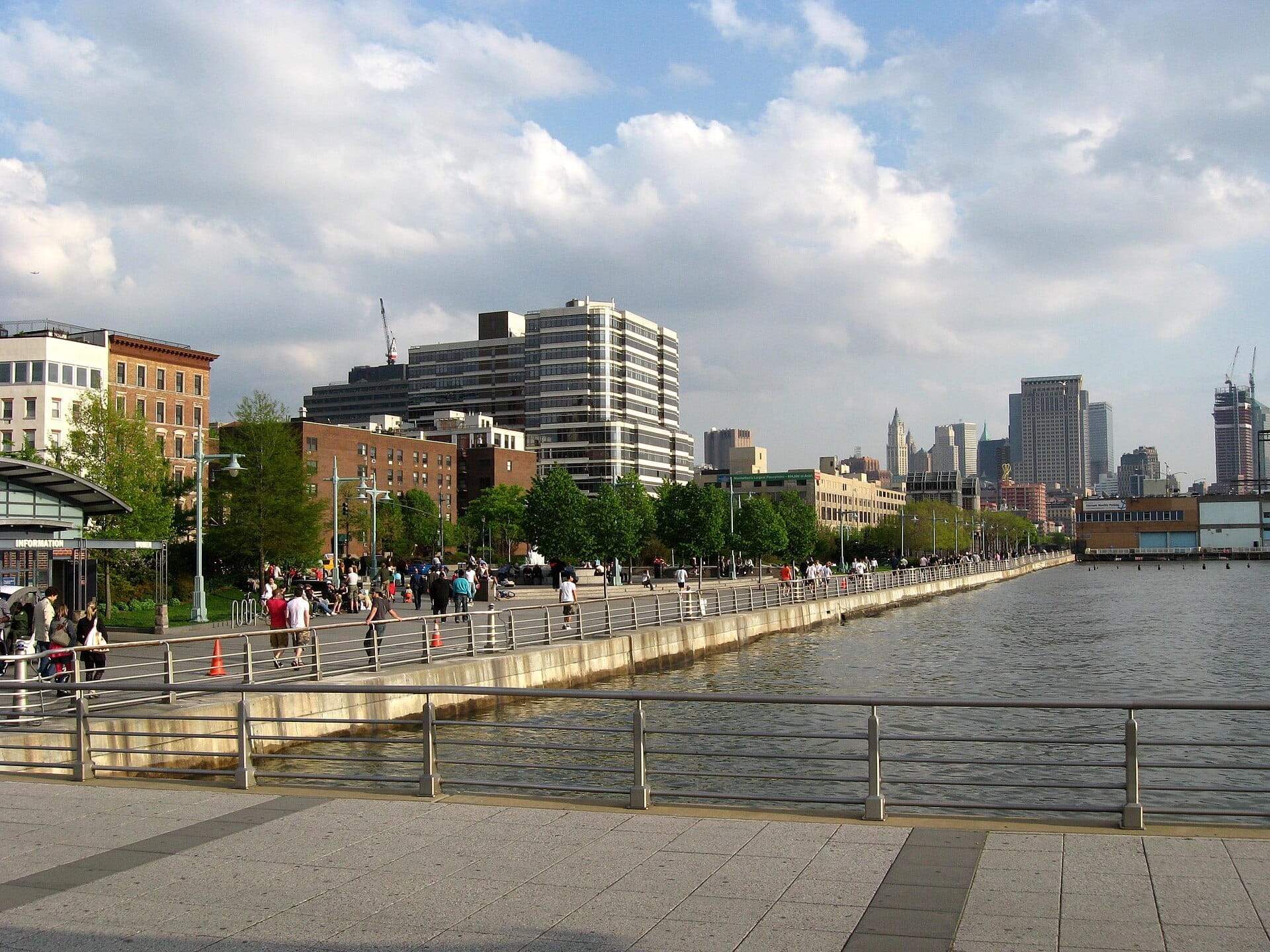 Le persone passeggiano lungo il lungofiume dell'Hudson River Park, con alberi, acqua e i grattacieli di Manhattan sotto un cielo parzialmente nuvoloso.