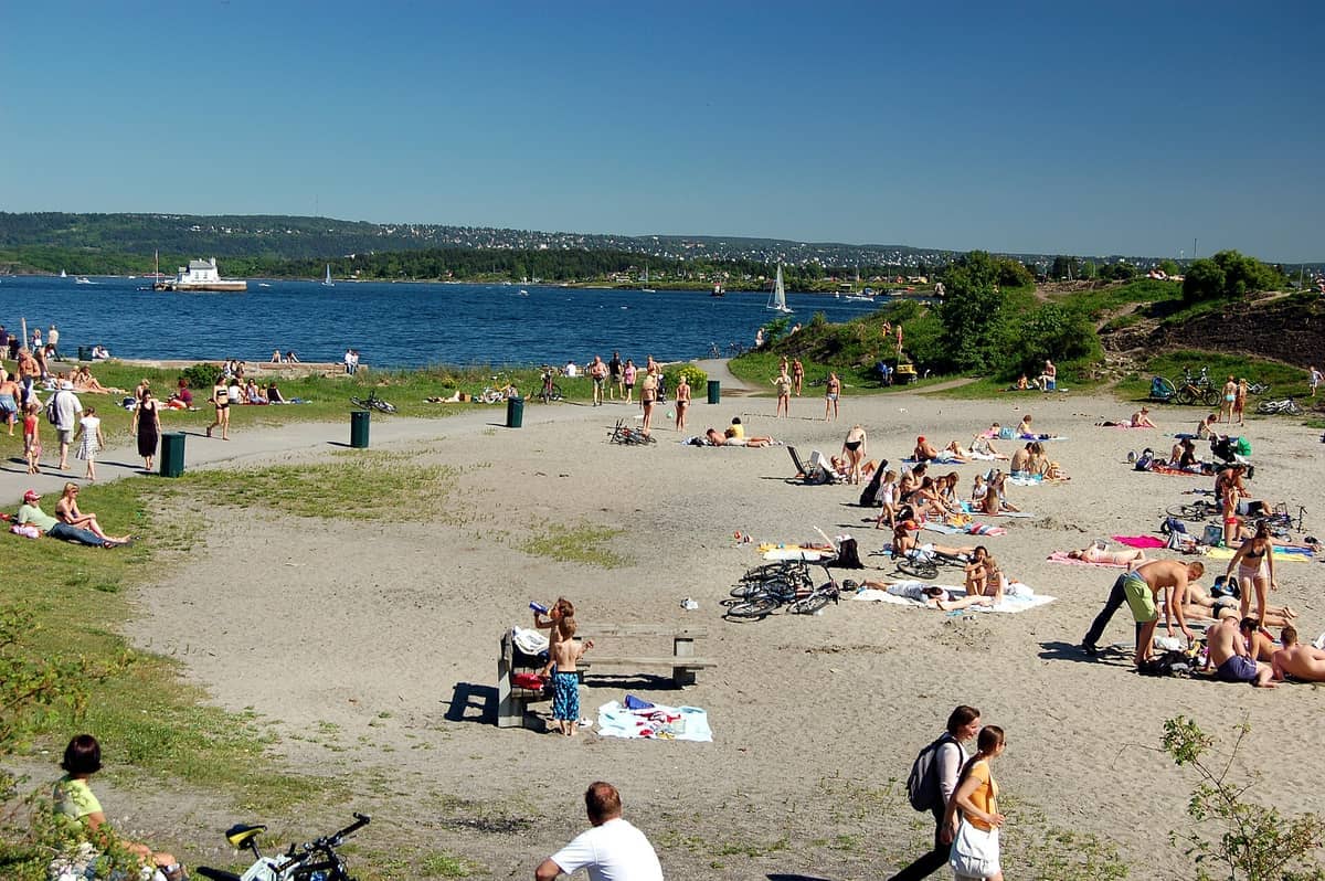 ผู้คนอาบแดดและพักผ่อนบนชายหาดทรายของ Huk Beach พร้อมพื้นที่สีเขียวและ Oslofjord ที่มองเห็นได้ภายใต้ท้องฟ้าสีฟ้าใส