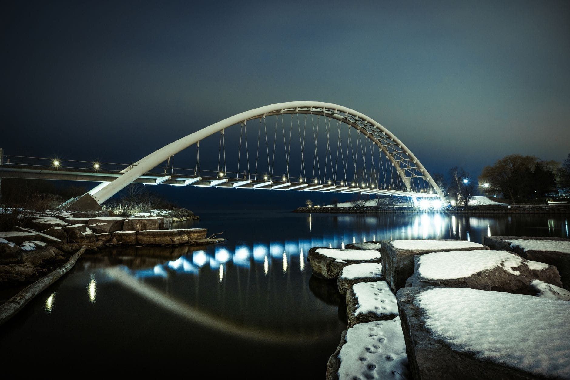 วิวกลางคืนของสะพานโค้ง Humber Bay ที่ส่องไฟสะท้อนผิวน้ำเงียบสงบในแม่น้ำ Humber พร้อมก้อนหิมะตามโขดหินริมฝั่ง