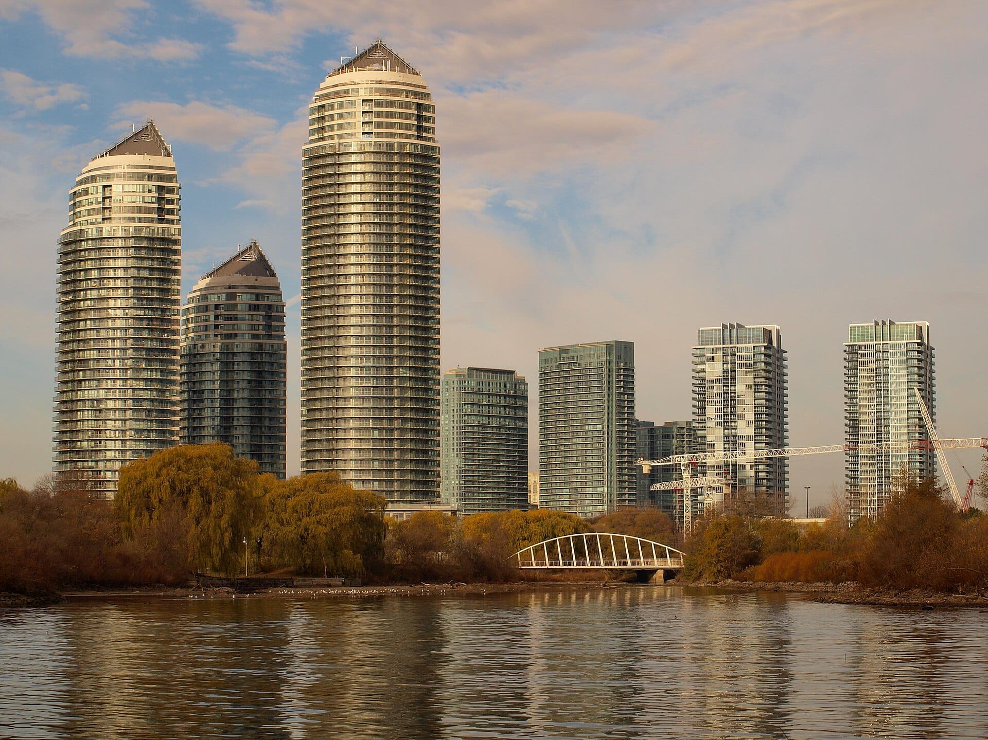 คอนโดมิเนียมสูงสมัยใหม่และสะพานคนเดินที่มองเห็นจากริมทะเลสาบใน Humber Bay Park พร้อมต้นไม้สีทองและน้ำนิ่งในเบื้องหน้า