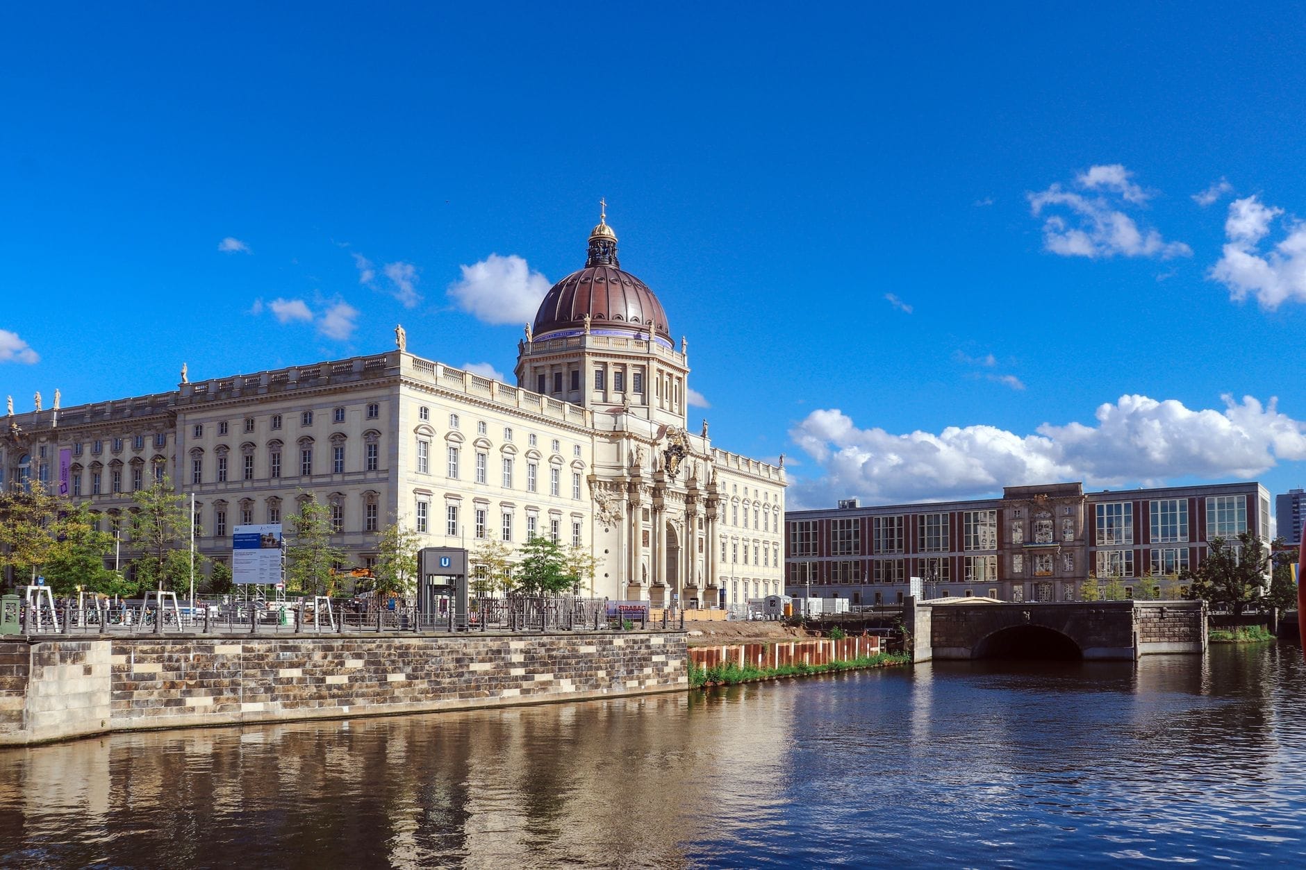 L'Humboldt Forum di Berlino, con la sua facciata barocca ricostruita e la grande cupola, visto dall'altra sponda del fiume sotto un cielo azzurro luminoso con qualche nuvola sparsa.
