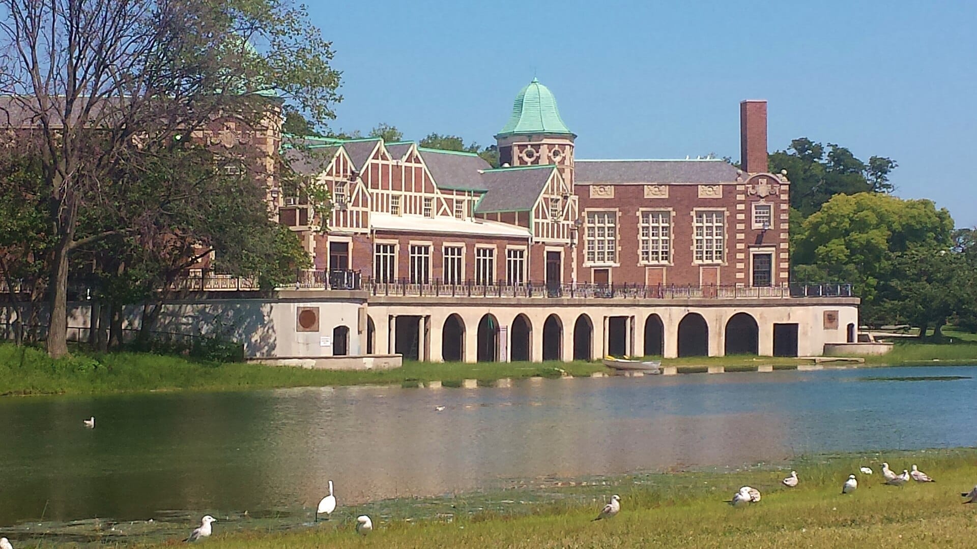 La storica rimessa per barche di Humboldt Park con colonnato ad archi si affaccia su una laguna tranquilla, circondata da vegetazione e uccelli in una giornata di sole.