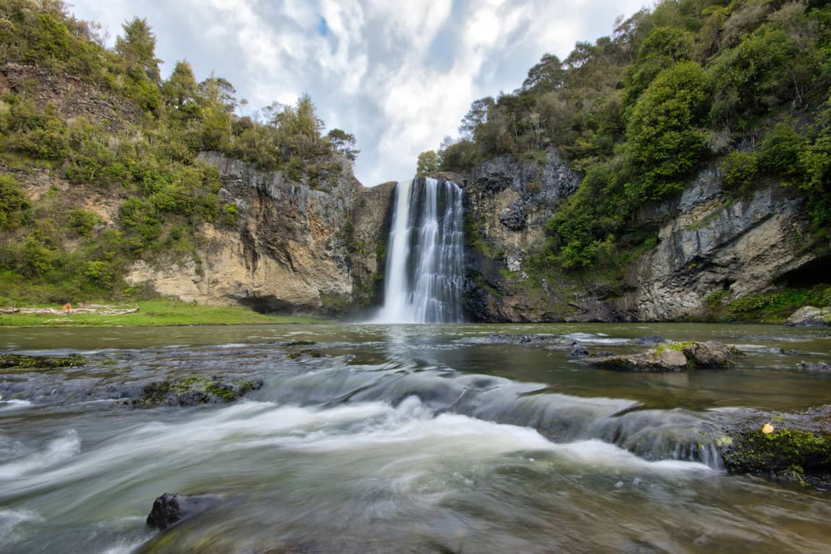 Panoramablick auf die Hūnua Falls, die in ein Becken stürzen, umgeben von üppigem Urwald und Felswänden unter teils bewölktem Himmel.