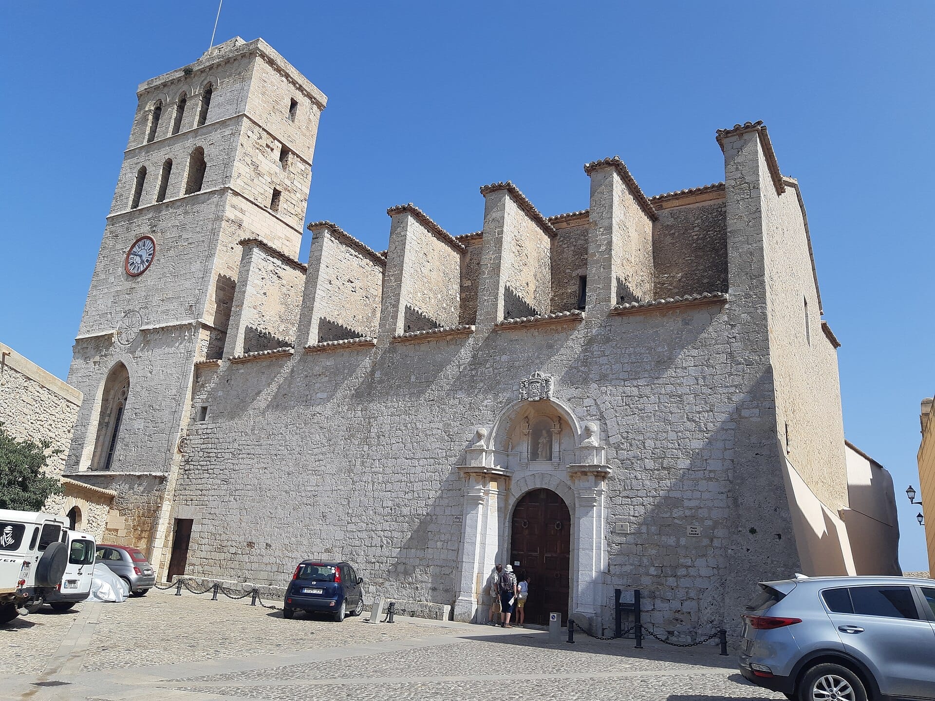 La facciata in pietra e il campanile della Cattedrale di Ibiza si innalzano contro un cielo blu limpido, con auto parcheggiate e visitatori vicino all'ingresso storico.