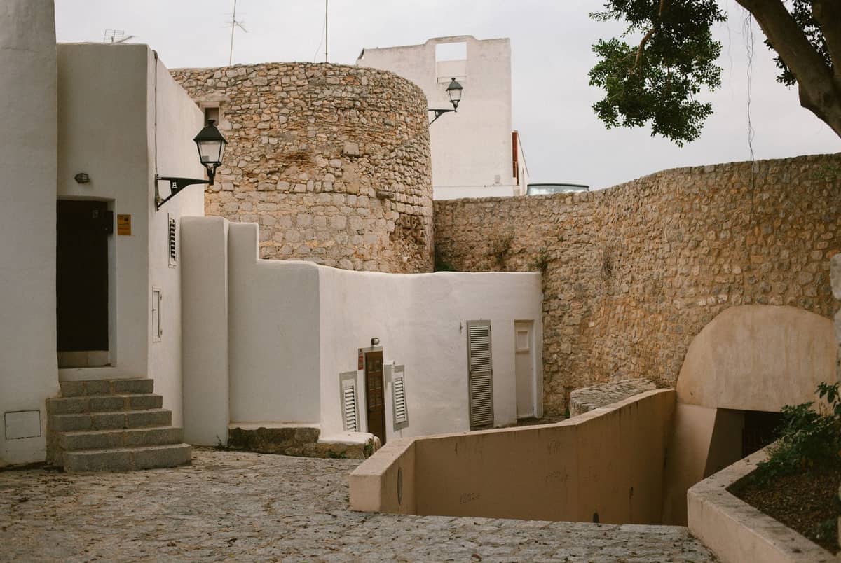 A cobblestone street winds through Dalt Vila, lined with whitewashed stone walls and historic buildings, showing a quiet area perfect for self-guided exploration.