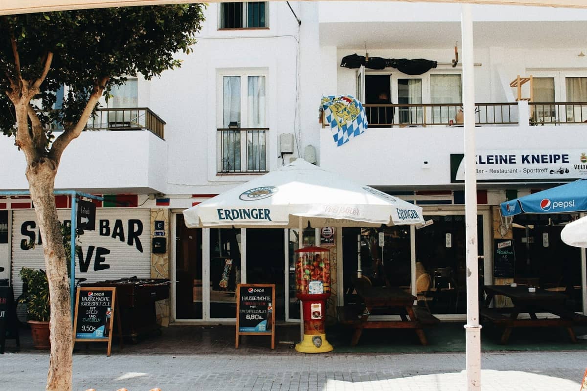 Street view of cafe and bar patios with umbrellas in a whitewashed Mediterranean building, suitable for eating or drinking in Ibiza.