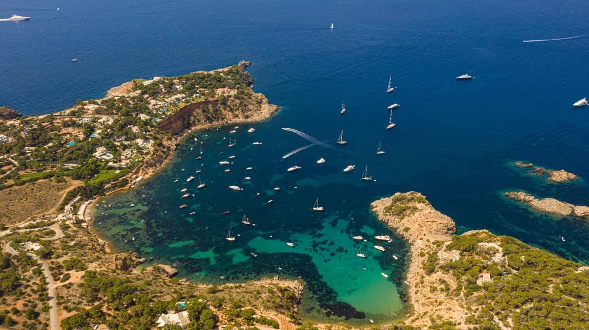 Aerial view of a bay in Ibiza with turquoise water, sailing boats, rocky coastline, and green hillsides with buildings.
