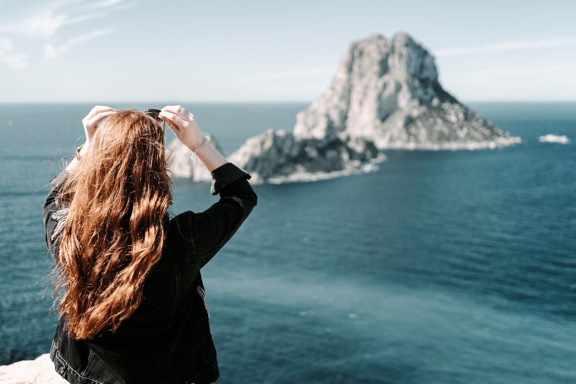 Uma mulher de cabelo comprido contempla o mar azul e a ilha de Es Vedrà ao largo da costa de Ibiza num dia de sol.