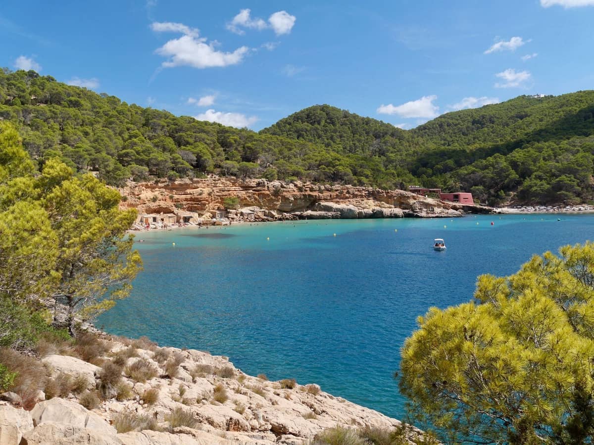 Quiet cove with turquoise water, rocky shoreline, and green pine hills, a boat anchored peacefully, evoking Ibiza's unspoiled beaches.