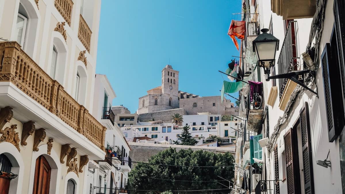 View up a narrow street in Ibiza old town, with historic stone buildings and the cathedral atop the hill under clear blue sky.