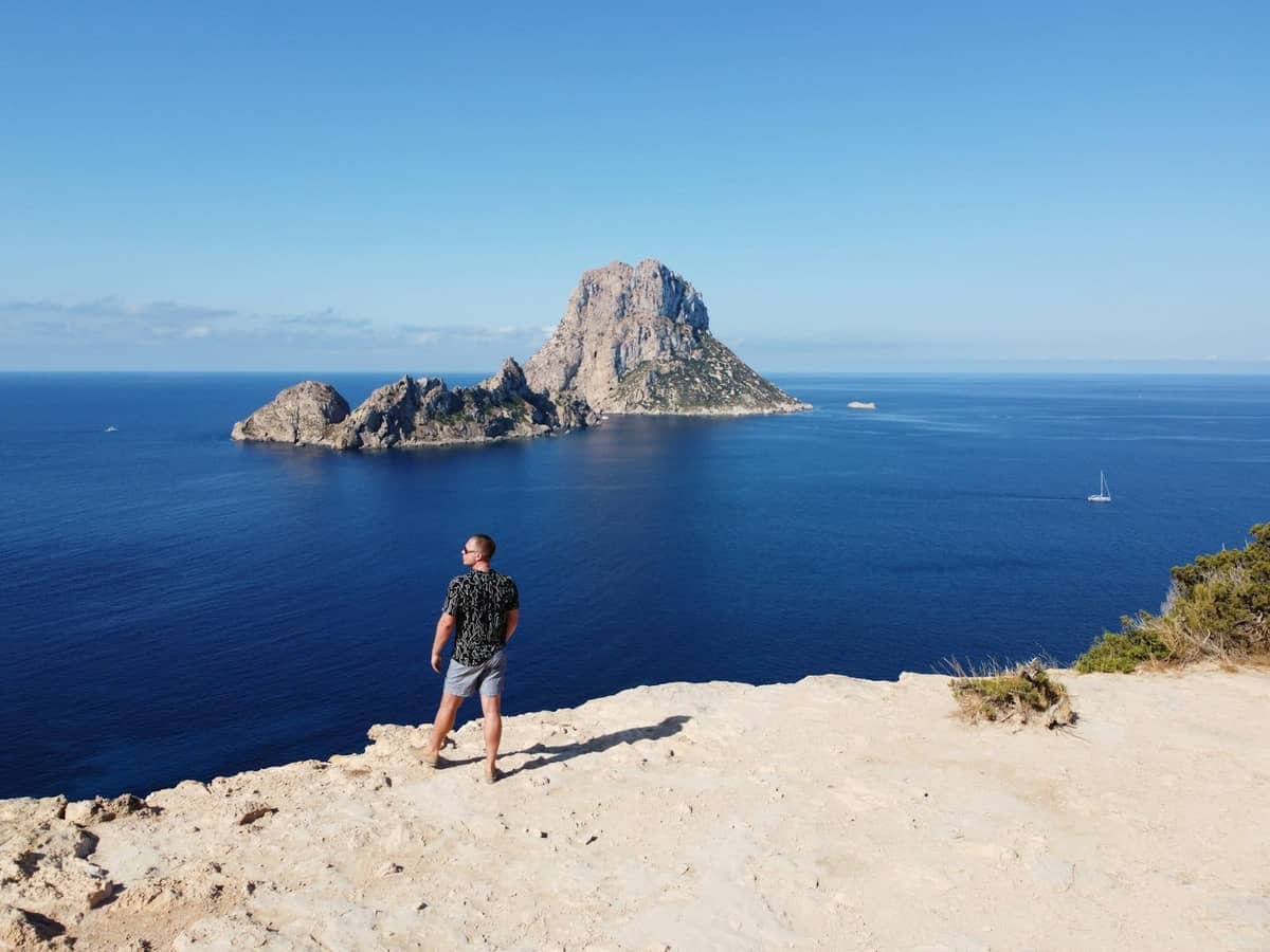 Hiker standing on a rocky summit overlooking deep blue sea and dramatic island cliff, clear sky, iconic Ibiza coastal scenery.