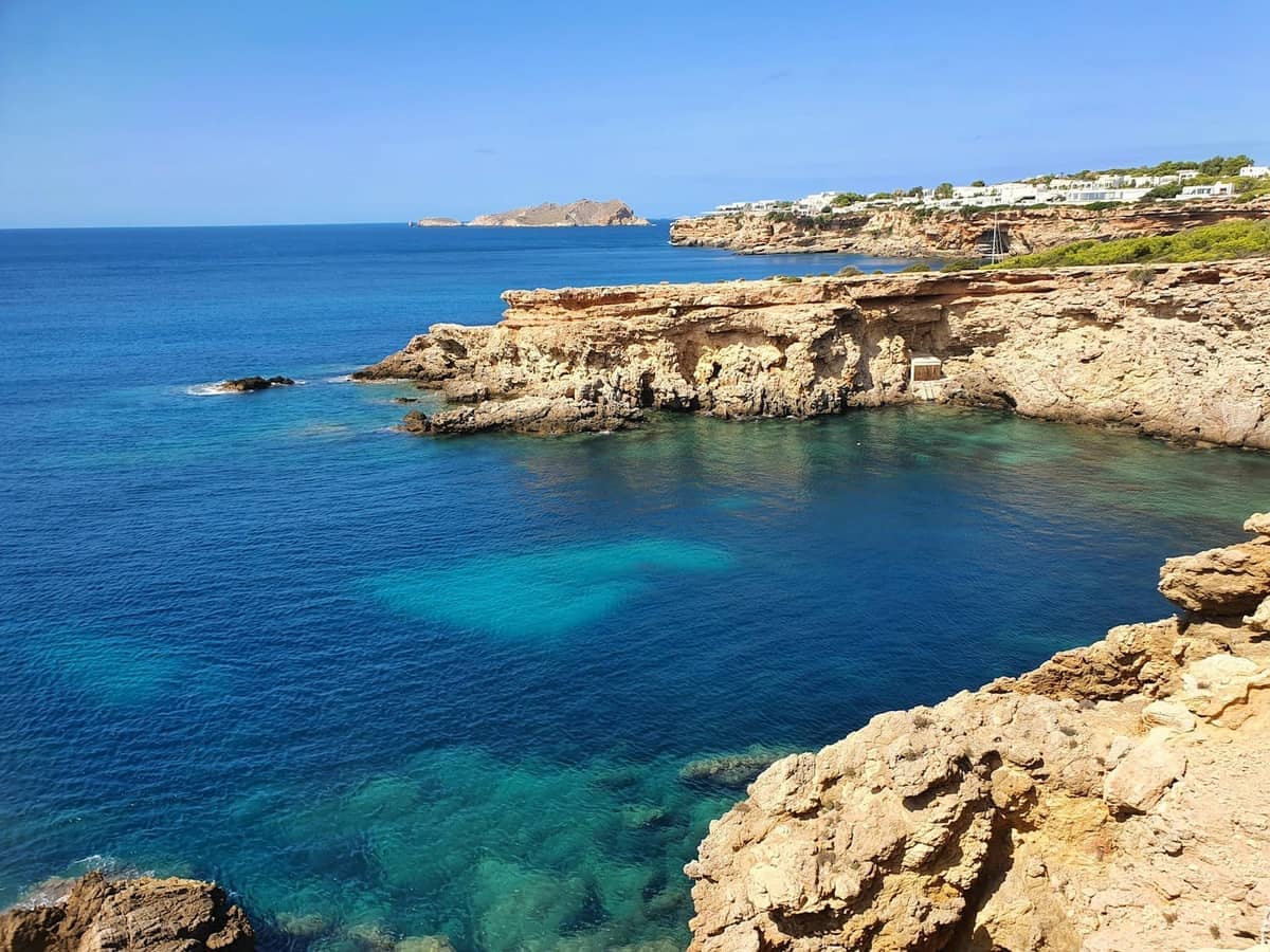 Rocky cliffs and clear turquoise sea along Ibiza's coastline, with distant white buildings and dramatic headlands under a bright blue sky.