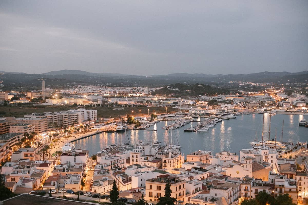 Aerial evening view of Ibiza Town harbour and marina, with white buildings and boats under city lights.
