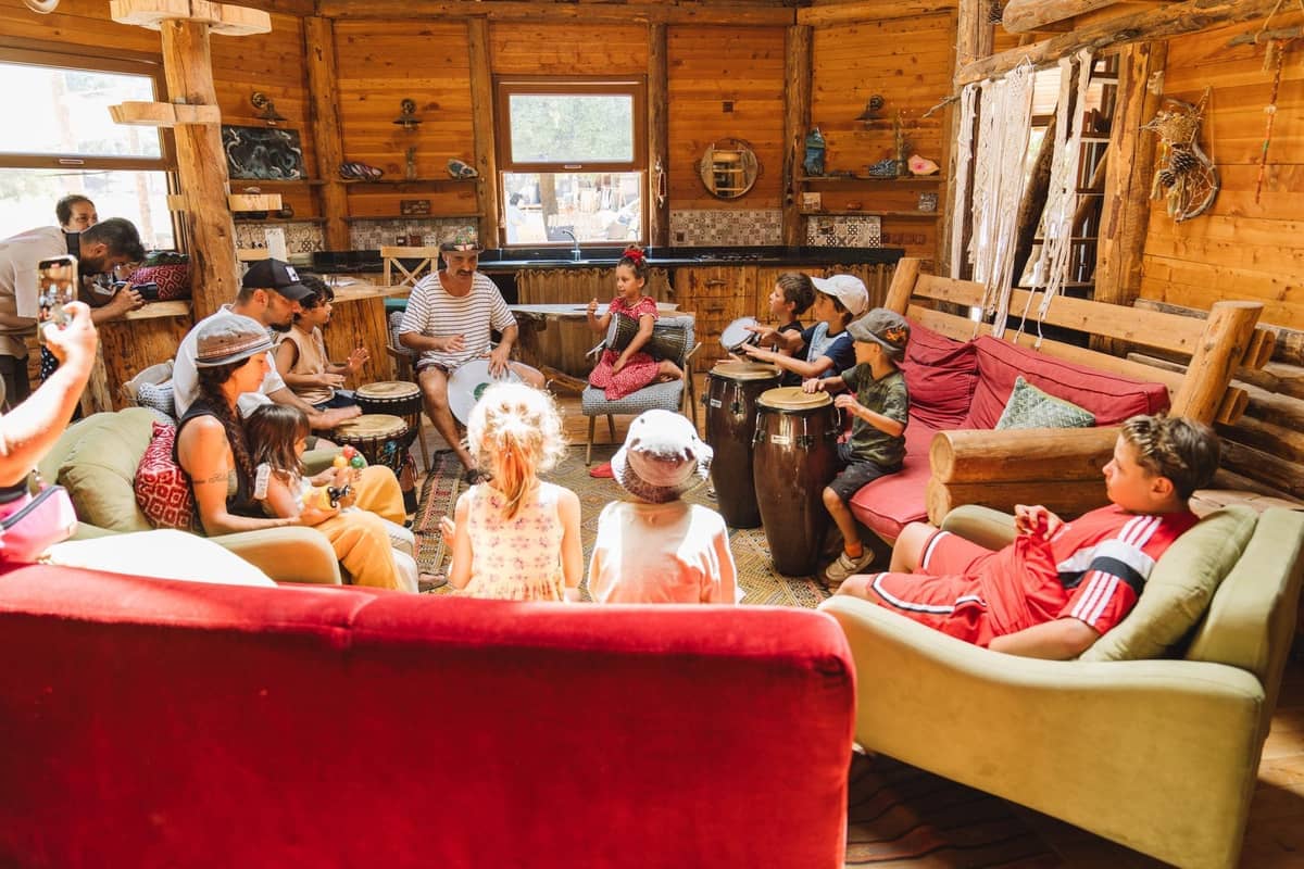 A group of people and children sitting in a circle inside a rustic wooden cabin, participating in a drum circle.