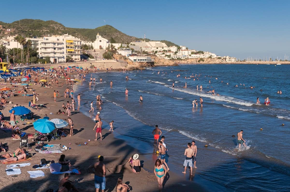 Busy Ibiza beach scene in late summer with sunbathers, swimmers, and people enjoying the water, lined by buildings and hills in the background.