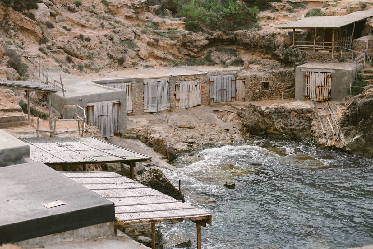 Empty and shuttered boathouses along a rocky Ibiza coastline, with closed doors and deserted appearance.