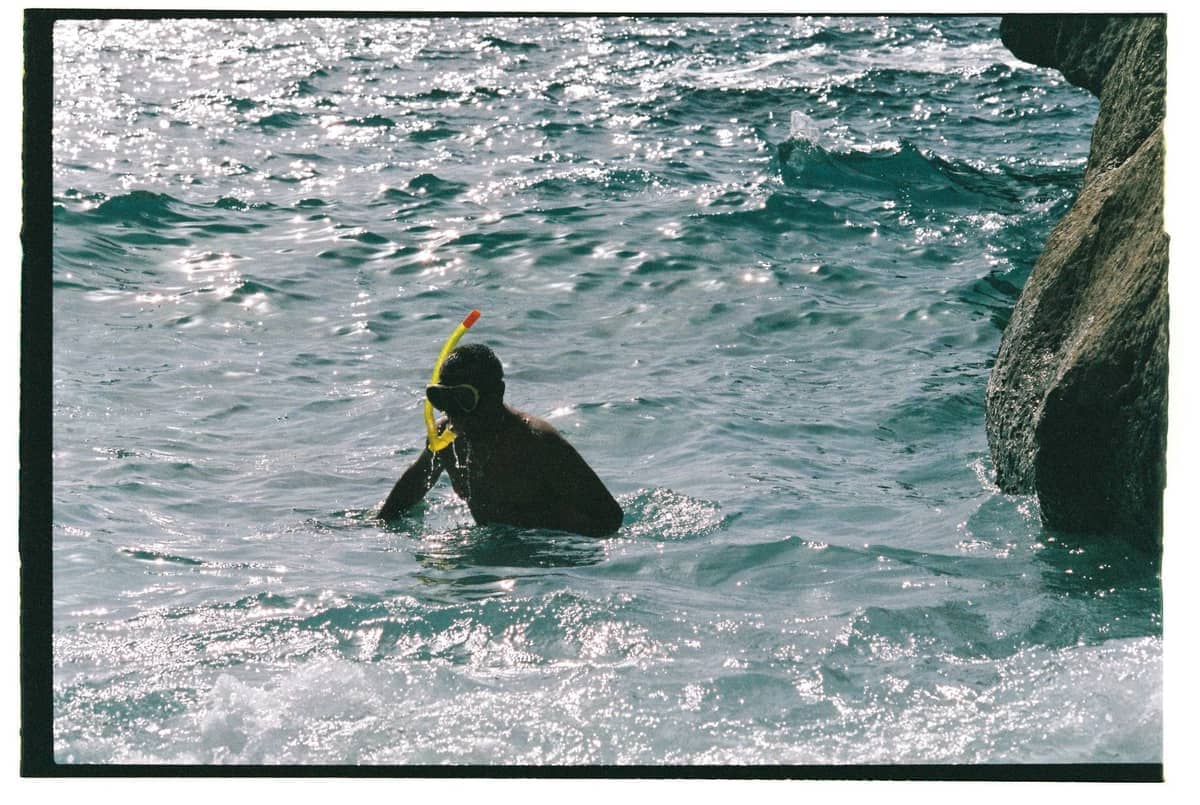 Person in snorkel gear swimming near rocky coast with clear water and sunlight sparkling on the sea