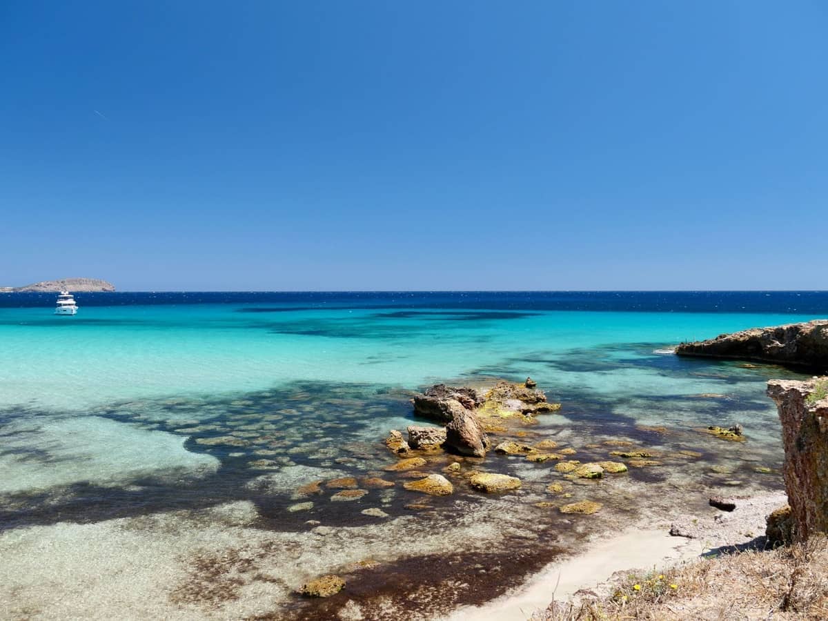 Clear turquoise water and rocky outcrops on a sunny beach, with a distant boat and islets off the western Ibiza coast.