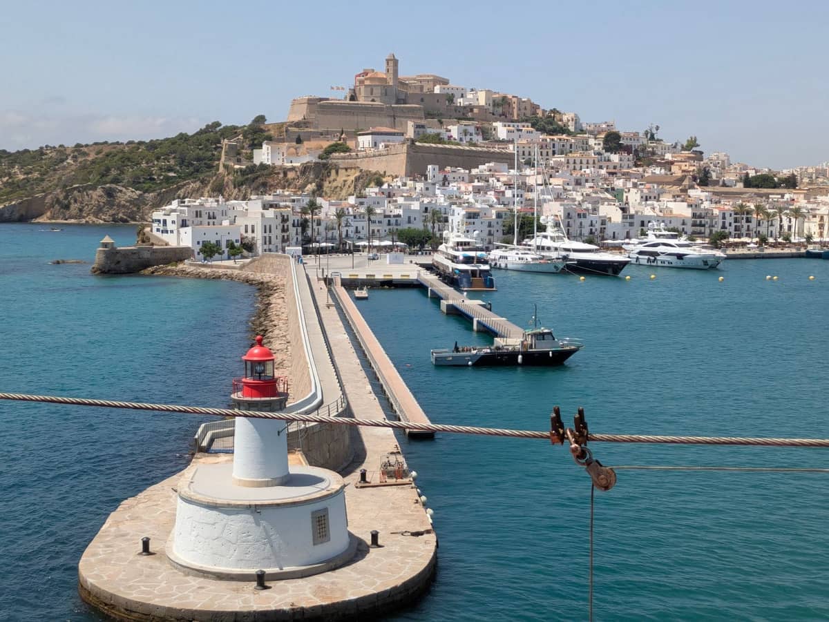Ibiza Town port with a red lighthouse and marina, historic old town buildings and fortress on a hill under clear sky.