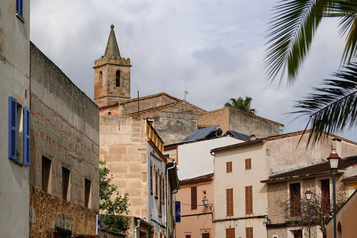 View of a Mediterranean village with whitewashed buildings and a church tower, surrounded by palm trees and narrow streets.
