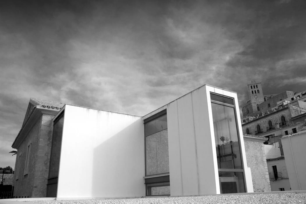 Vista esterna del Museo d'Arte Contemporanea di Ibiza (MACE) con architettura moderna in vetro accanto a edifici storici in pietra sotto un cielo nuvoloso spettacolare.