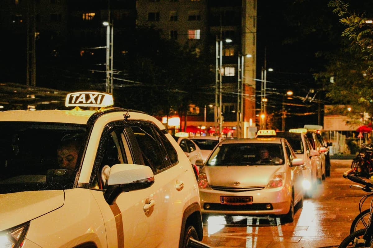 A line of white taxis waiting at night on a wet, well-lit city street with buildings and streetlights visible in the background.