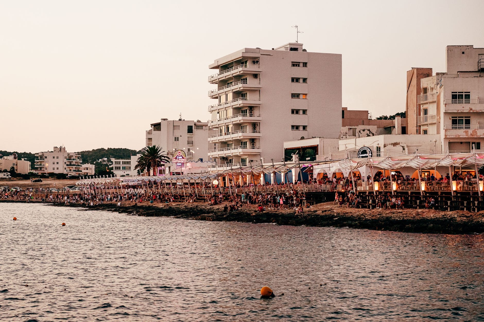 Fileira animada de bares e restaurantes à beira-mar lotados de pessoas ao entardecer, com prédios modernos e palmeiras ao fundo, capturando a atmosfera noturna de Ibiza.