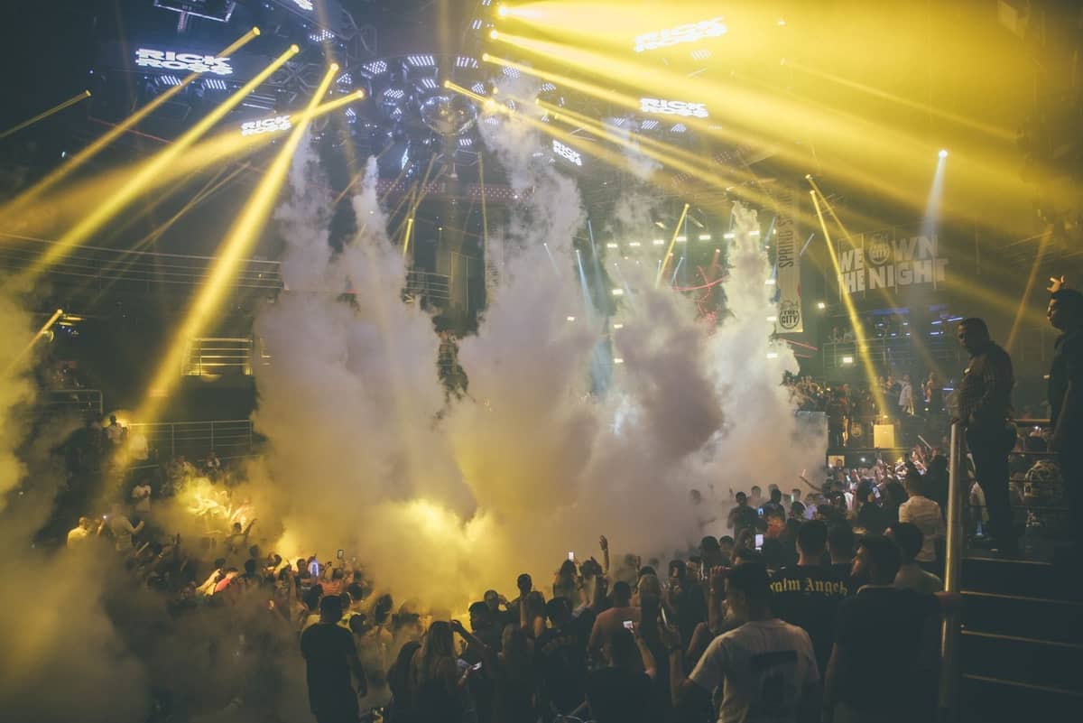 Packed nightclub with bright yellow lights, dramatic smoke effects, and energetic crowd dancing on the main floor at night.